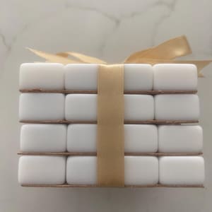Pu&ograve; includere: Una pila di oggetti rettangolari bianchi, forse sottobicchieri, legati con un nastro dorato. Gli oggetti sono disposti in quattro strati, ciascuno con quattro pezzi. Il nastro &egrave; legato con un fiocco in alto.