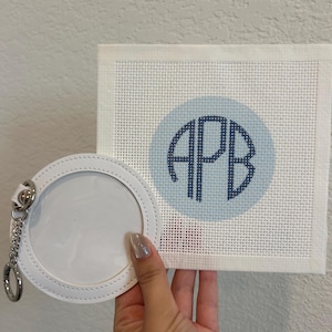 May include: A square white needlepoint canvas featuring a light blue circle with the dark blue monogram letters APB stitched in the center, accompanied by a circular white leather-like keychain with a clear plastic window and a silver metal ring.