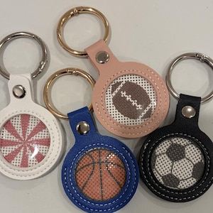 May include: Four circular keychains with gold-tone metal rings. The white keychain features a pink starburst pattern, the blue one shows a basketball, the peach one displays an American football, and the black one features a soccer ball pattern.