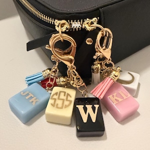 May include: A black structured case with a gold zipper featuring two gold-toned lobster clasp keychains. The keychains are adorned with colorful tassels and rectangular charms in light blue, cream, black, and pink, each displaying gold letters JTK, SSS, W, and KL.
