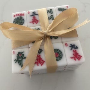Pu&ograve; includere: Un set di tessere di Mahjong bianche legate con un nastro dorato. Le tessere presentano simboli e numeri in rosso, verde e nero. Una tessera indica "JOKER".