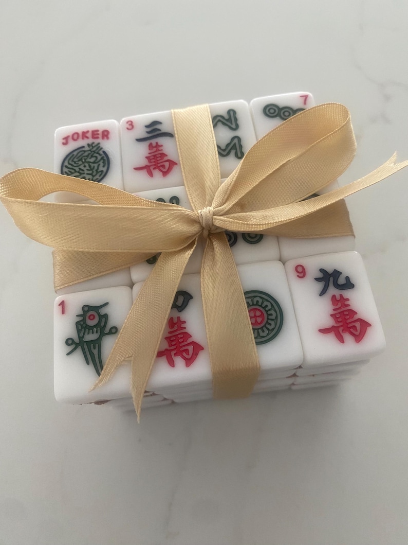 Pu&ograve; includere: Un set di tessere di Mahjong bianche legate con un nastro dorato. Le tessere presentano simboli e numeri in rosso, verde e nero. Una tessera indica "JOKER".