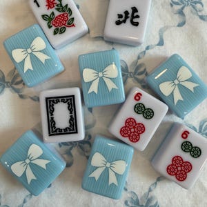 May include: A collection of Mahjong tiles. The tiles are white, light blue, and feature various symbols, numbers, and characters. Some tiles have a white bow design on a light blue background. Other tiles have floral and number designs.