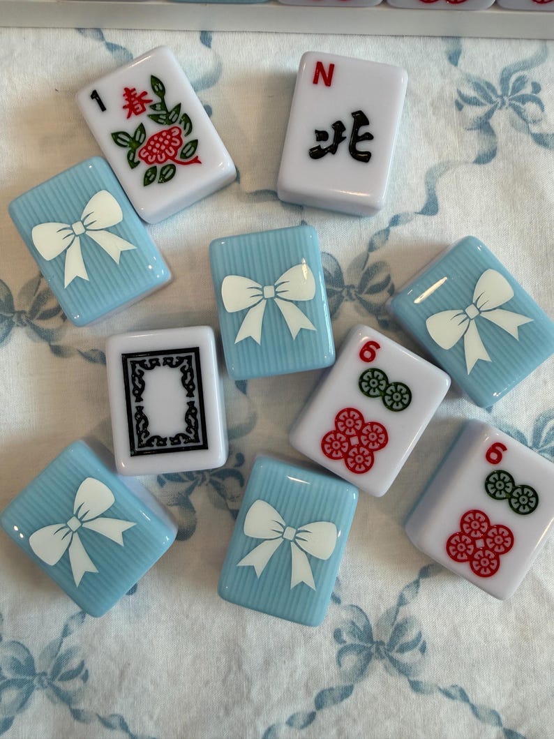 May include: A collection of Mahjong tiles. The tiles are white, light blue, and feature various symbols, numbers, and characters. Some tiles have a white bow design on a light blue background. Other tiles have floral and number designs.