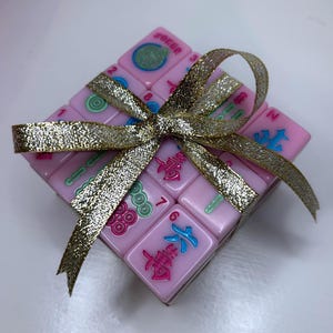 Pu&ograve; includere: Un quadrato di tessere rosa del Mahjong, legato con un nastro dorato glitterato. Le tessere presentano simboli e numeri colorati. La parola "Joker" &egrave; visibile su una tessera. Il nastro &egrave; legato a fiocco.