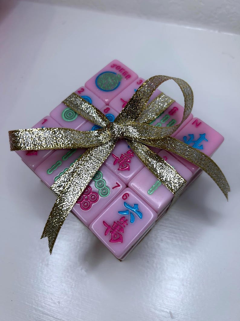 Pu&ograve; includere: Un quadrato di tessere rosa del Mahjong, legato con un nastro dorato glitterato. Le tessere presentano simboli e numeri colorati. La parola "Joker" &egrave; visibile su una tessera. Il nastro &egrave; legato a fiocco.