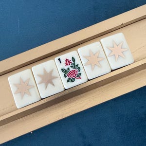 May include: Five rectangular mahjong tiles arranged in a row on a wooden rack. Four tiles feature a light pink eight-pointed star design on a white background. The center tile displays the number 1, a red flower, and green leaves with the character 春.