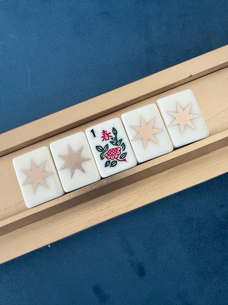 May include: Five rectangular mahjong tiles arranged in a row on a wooden rack. Four tiles feature a light pink eight-pointed star design on a white background. The center tile displays the number 1, a red flower, and green leaves with the character 春.