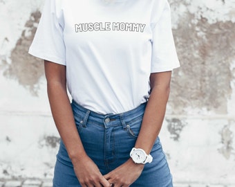 May include: A white t-shirt with the text "MUSCLE MOMMY" printed on the front. The shirt is being worn by a person with blonde hair and blue jeans.