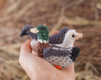 May include: Three crocheted stuffed ducks held in a hand. One duck is grey and white, one is grey and brown, and one is green, brown, and white.