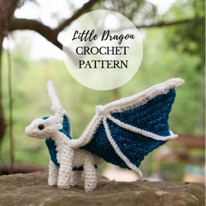 May include: A crocheted dragon with white and blue details, including wings and a small head. The dragon is standing on a rock. The image includes the text "Little Dragon CROCHET PATTERN".