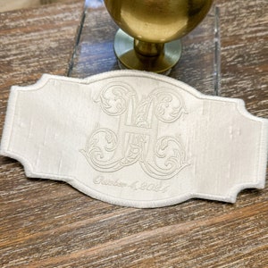 May include: Ivory-colored embroidered label with a decorative monogram and the date "October 4, 2024". The label has a scalloped edge and is set against a wooden surface.