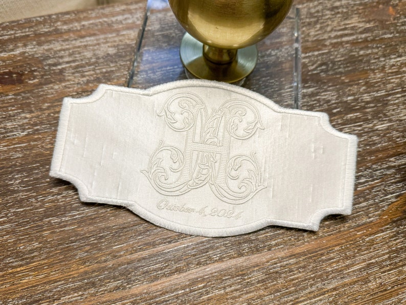 May include: Ivory-colored embroidered label with a decorative monogram and the date "October 4, 2024". The label has a scalloped edge and is set against a wooden surface.