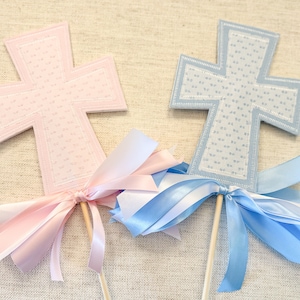 May include: Two cross-shaped decorations, one pink and one blue, with floral patterns and ribbon bows. Each cross is attached to a wooden stick. The pink cross has pink and white ribbons, while the blue cross has blue and white ribbons.
