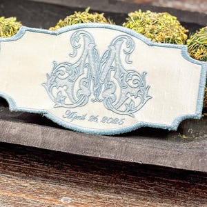May include: An embroidered ivory-colored rectangular patch with a light blue border. The patch features a large, ornate light blue letter "M" and the date "April 26, 2025" embroidered in a script font. The patch is placed on a wooden surface.