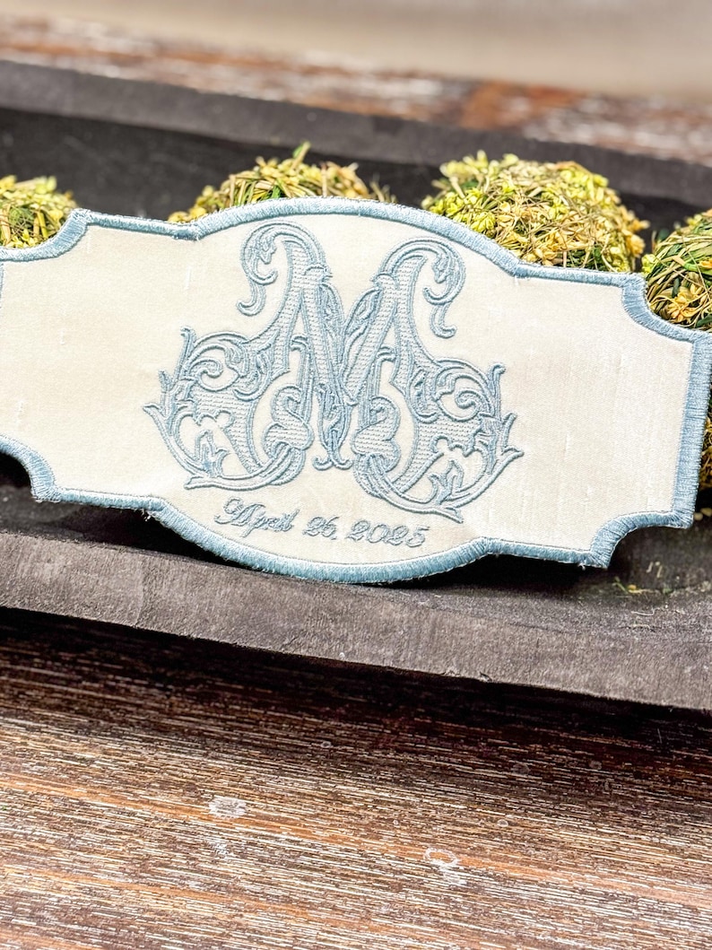 May include: An embroidered ivory-colored rectangular patch with a light blue border. The patch features a large, ornate light blue letter "M" and the date "April 26, 2025" embroidered in a script font. The patch is placed on a wooden surface.