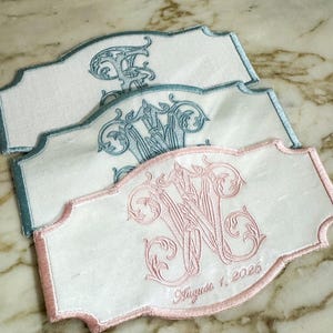 May include: Three embroidered white fabric labels with scalloped edges. One label features a pink monogram and the date "August 1, 2026." The other two labels have blue monograms. The embroidery is ornate and detailed.