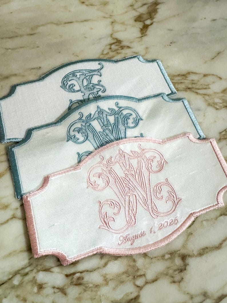 May include: Three embroidered white fabric labels with scalloped edges. One label features a pink monogram and the date "August 1, 2026." The other two labels have blue monograms. The embroidery is ornate and detailed.