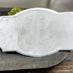 May include: White embroidered decorative piece with an ornate, stylized letter "J" in the center. The piece has a rectangular shape with a scalloped edge. The embroidery is detailed and raised, creating a textured effect.