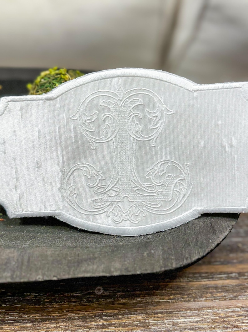 May include: White embroidered decorative piece with an ornate, stylized letter "J" in the center. The piece has a rectangular shape with a scalloped edge. The embroidery is detailed and raised, creating a textured effect.
