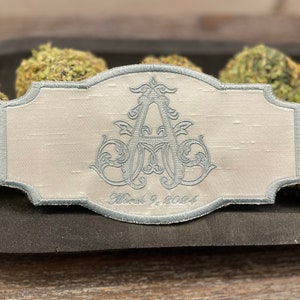 May include: A light gray embroidered patch with a decorative border. The patch features a stylized monogram in light blue thread, with the date "March 9, 2024" embroidered below. The patch is set against a textured background.