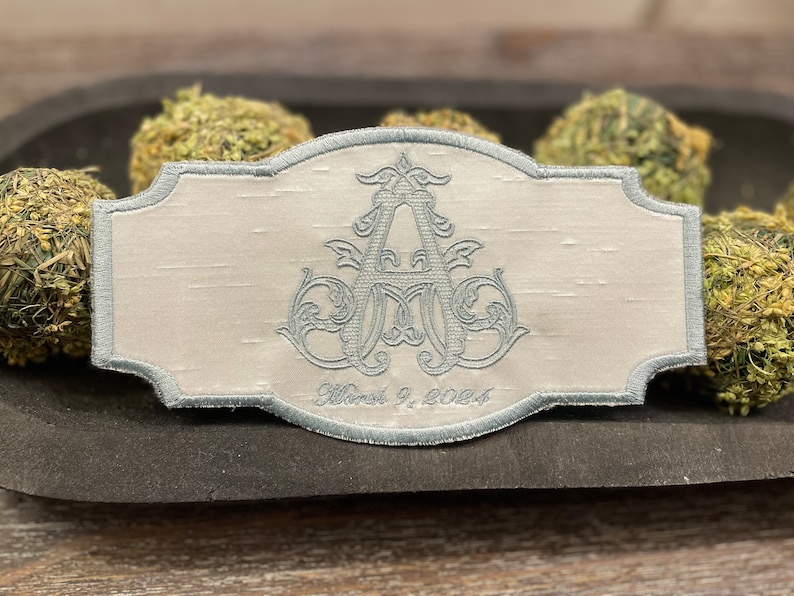 May include: A light gray embroidered patch with a decorative border. The patch features a stylized monogram in light blue thread, with the date "March 9, 2024" embroidered below. The patch is set against a textured background.