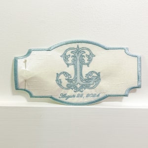 May include: An embroidered ivory-colored patch with a light blue border and a decorative initial "J". The patch has the date "August 22, 2024" embroidered in a script font. A pin is attached to the patch.
