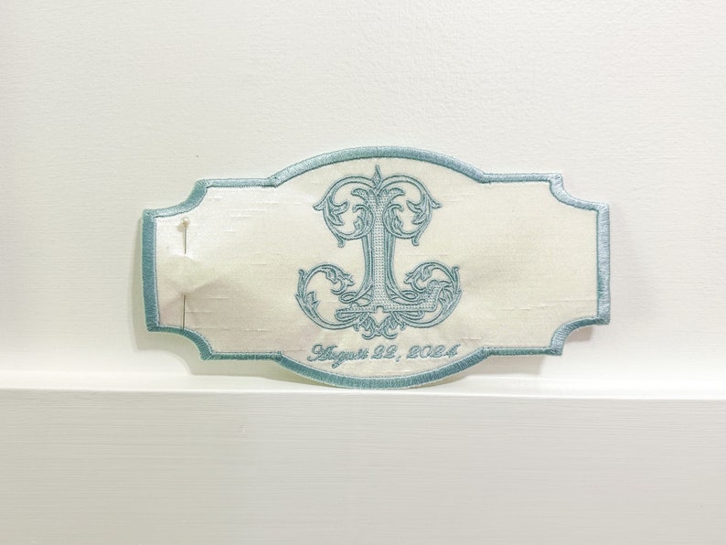 May include: An embroidered ivory-colored patch with a light blue border and a decorative initial "J". The patch has the date "August 22, 2024" embroidered in a script font. A pin is attached to the patch.