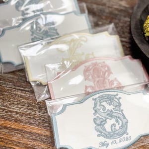 May include: Embroidered white labels with ornate designs in blue, yellow, and pink, packaged in clear plastic. The labels feature a decorative border and a monogrammed letter with the date "May 10, 2025".