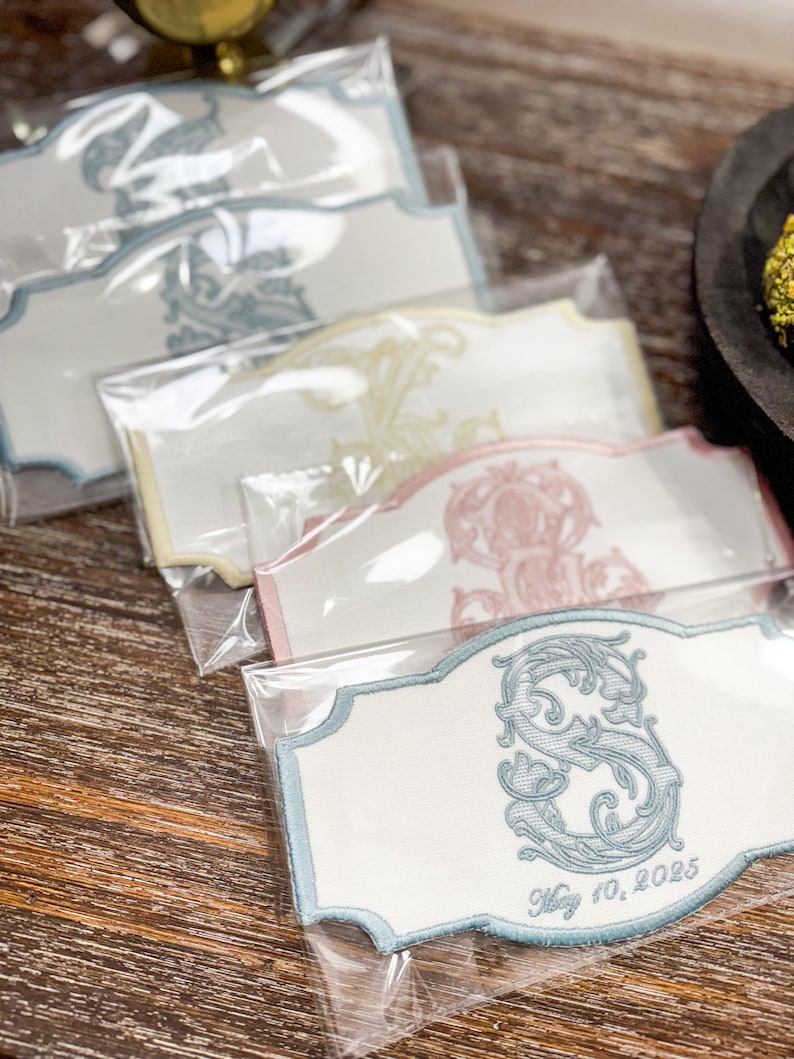 May include: Embroidered white labels with ornate designs in blue, yellow, and pink, packaged in clear plastic. The labels feature a decorative border and a monogrammed letter with the date "May 10, 2025".