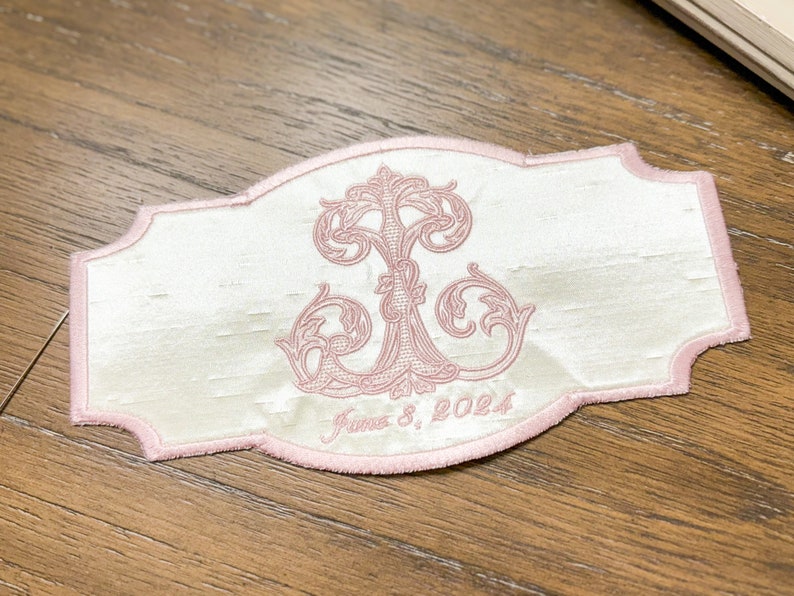 May include: A white embroidered patch with a pink border and a pink floral design. The patch has the text "June 8, 2024" embroidered in pink. The patch is rectangular with a decorative edge.
