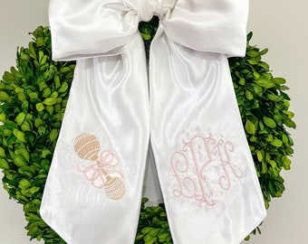 May include: A large white satin bow with long streamers, adorned with a light pink monogram and a rattle embroidery. The bow is set against a backdrop of a vibrant green boxwood wreath, creating a classic and elegant look.