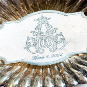 May include: A white rectangular embroidered patch with a light blue border and a light blue monogram. The monogram is an ornate "A" with decorative flourishes. The date "March 9, 2024" is embroidered below the monogram.