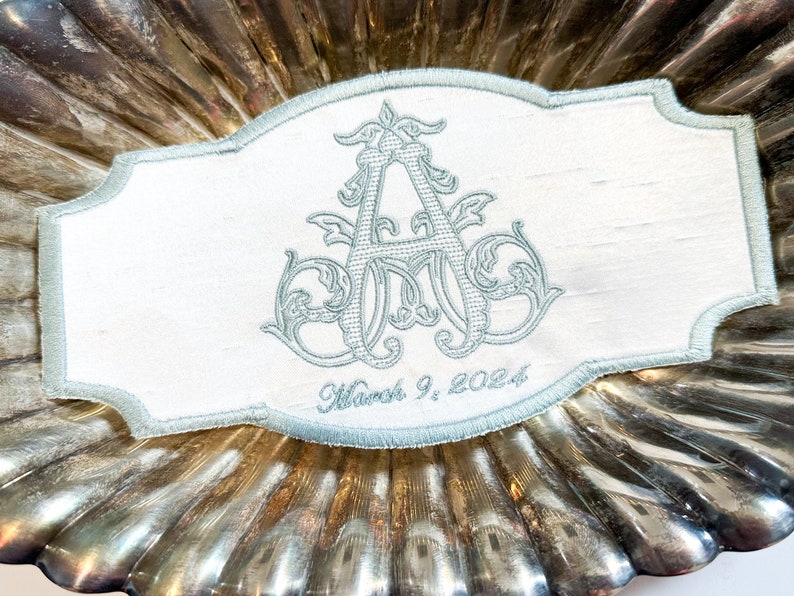 May include: A white rectangular embroidered patch with a light blue border and a light blue monogram. The monogram is an ornate "A" with decorative flourishes. The date "March 9, 2024" is embroidered below the monogram.