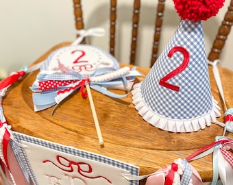 May include: A festive birthday set featuring a blue and white gingham high chair banner with embroidered initials and a red bow. Includes a matching party hat with a red pom-pom and the number 2, and a cake topper.