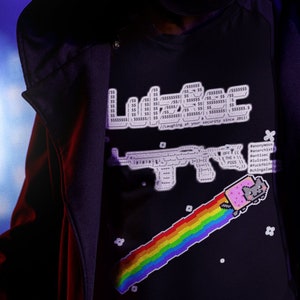 May include: A person wearing a black hooded jacket and a black t-shirt with a rainbow and a cat graphic. The t-shirt has the text "L33T" printed on it.