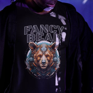 Peut inclure: Une personne portant un t-shirt noir à manches longues avec le texte "FANCY BEAR" et un graphique d'un ours avec un design géométrique. La personne porte des lunettes roses et noires et une ceinture noire.