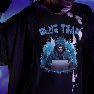Può includere: Una persona indossa una maglietta nera con una grafica blu di una figura con il cappuccio che usa un computer portatile. Il testo "BLUE TEAM" è stampato sulla maglietta in blu.