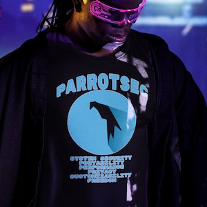 Op de afbeelding: Een persoon draagt een zwart T-shirt met een blauwe cirkel en een witte papegaai-afbeelding. Het shirt zegt "Parrotsec" en "System Security,  Post-Scarcity,  Privacy,  Open Source,  Bracket Left,  Freedom".