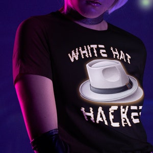 Könnte beinhalten: Eine Frau trägt ein schwarzes T-Shirt mit dem Text "WHITE HAT HACKERS" und einer weißen Fedora-Grafik. Sie trägt lila Haare, schwarze Handschuhe und eine silberne Halskette.