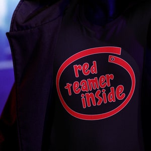 Puede incluir: Una persona que lleva una chaqueta negra con capucha y una camiseta negra con un gráfico circular rojo que dice "red teamer inside".