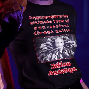 Peut inclure: Un t-shirt noir avec un imprimé graphique rouge représentant un homme avec de multiples mains tenant des armes à feu pointées sur sa tête. Le texte sur le t-shirt est "Cryptography is the ultimate form of non-violent direct action." et "Julian Assange".
