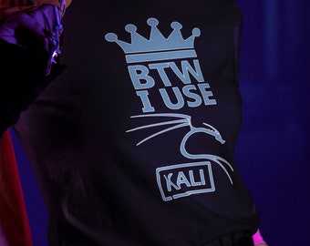 Op de afbeelding: Een persoon draagt een zwart T-shirt met een blauwe kroon en de tekst "BTW I USE KALI" erop gedrukt. De persoon heeft paars haar en draagt een gezichtsmasker.