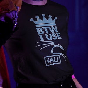 May include: A person wearing a black t-shirt with a blue crown and the text "BTW I USE KALI" printed on it. The person has purple hair and is wearing a face mask.