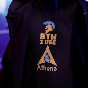 Puede incluir: Una chaqueta negra con capucha y una camiseta negra debajo. La camiseta tiene un gráfico dorado y azul con el texto "BTW I USE Athena".