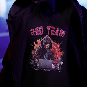Puede incluir: Una camiseta negra con un gráfico rojo de una persona con capucha usando una computadora portátil. El texto "RED TEAM" está por encima del gráfico.