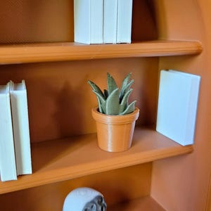 May include: A miniature orange arched bookshelf featuring three white books on the top shelf. The middle shelf holds two white books and a small green succulent plant in an orange pot. A white object is visible on the bottom shelf.
