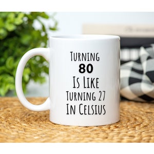 May include: White ceramic coffee mug with black text that reads "Turning 80 is like turning 27 in Celsius".