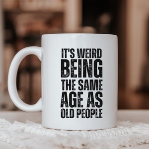 May include: White ceramic mug with black text that reads "It's weird being the same age as old people".
