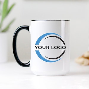 May include: White ceramic mug with a black rim and handle. The mug has a blue and black circle design with the text "YOUR LOGO" in black.